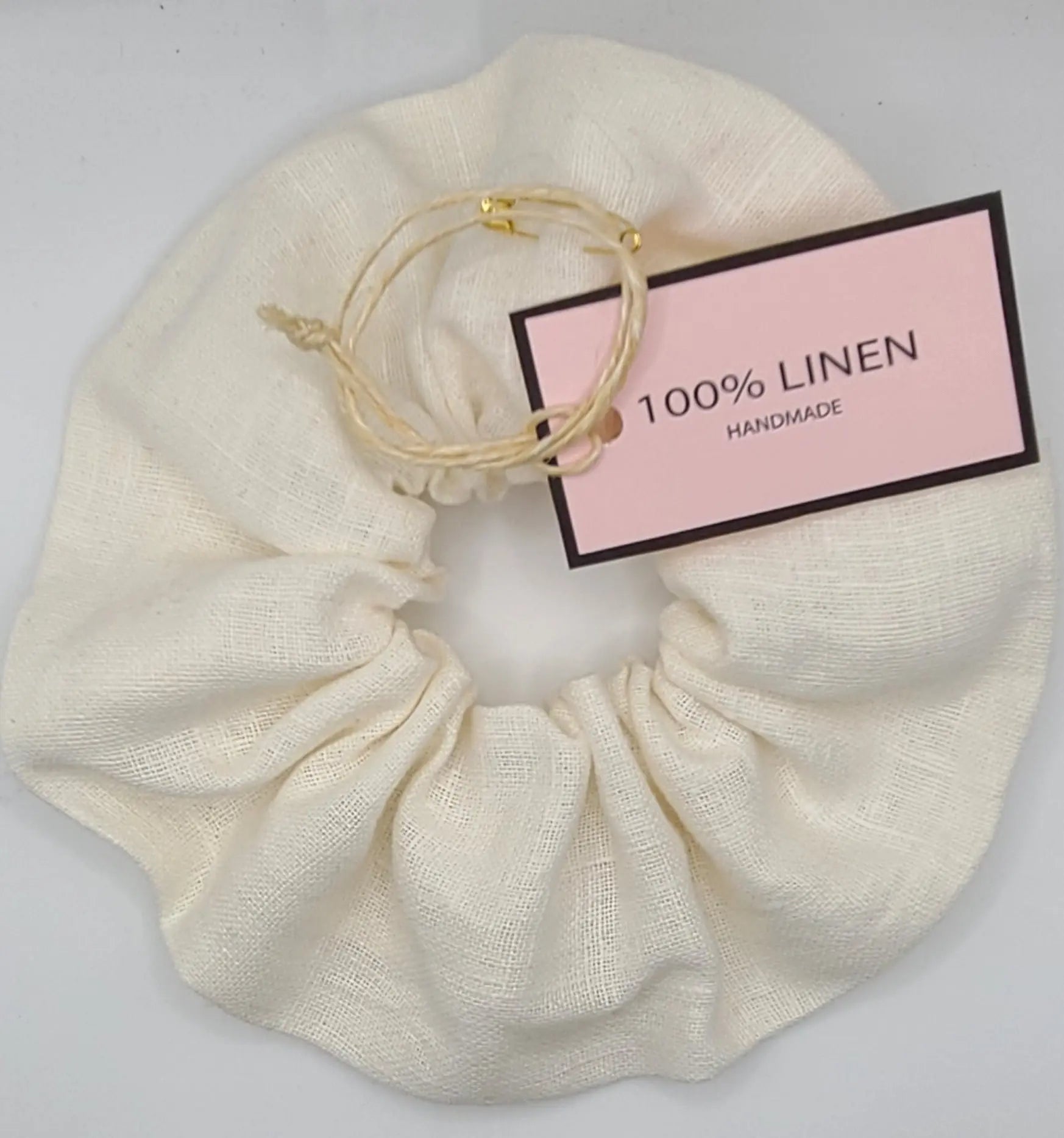 Cream-colored linen scrunchie with a gold hair tie and a label indicating '100% linen' and 'handmade' on a light gray background.